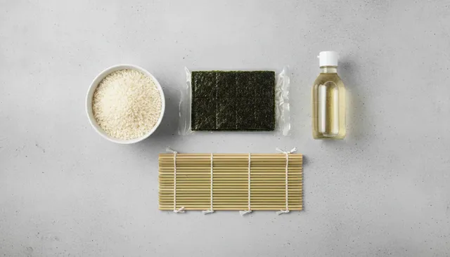 De basis-ingrediënten voor het maken van sushi van bovenaf gefotografeerd: een bamboematje, norivellen, een kom met sushirijst en een flesje rijstazijn.
